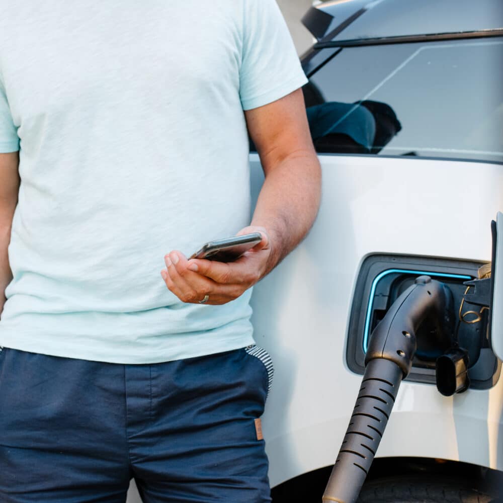 man on a smartphone by his electric vehicle charging at a station